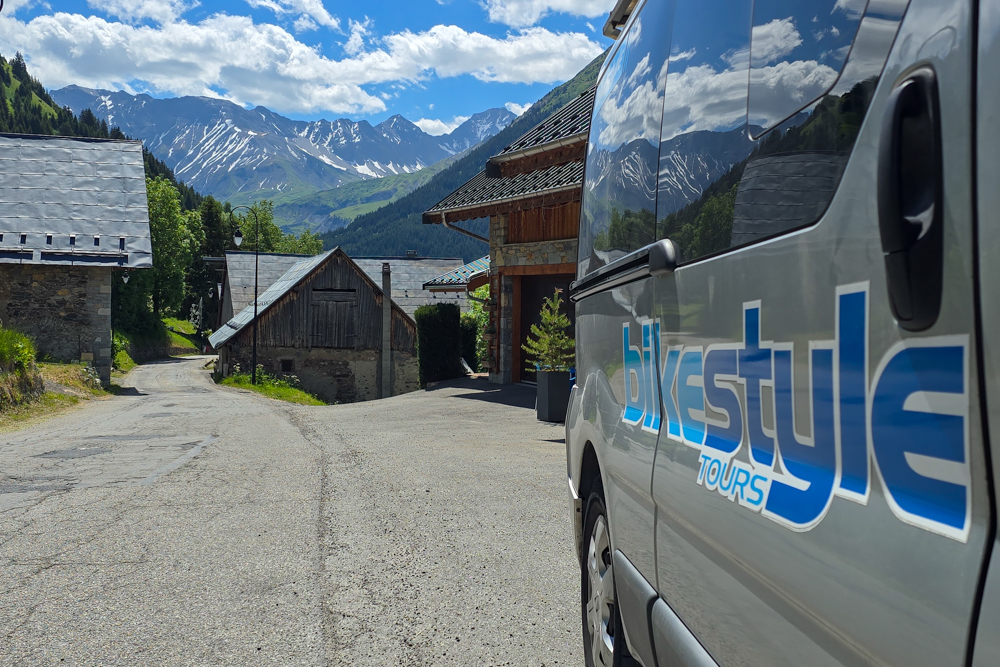 Tour van with blue branding parked along a mountain village street with wooden chalets and snow-capped peaks in the background.