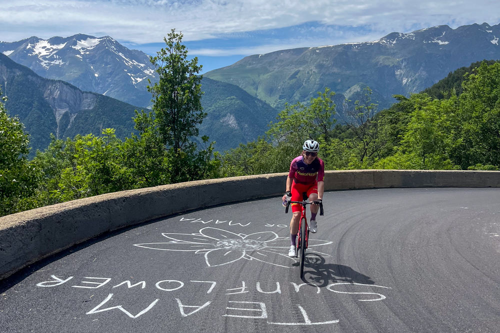 Female cyclist in pink and red kit rides a winding mountain road with chalk art on the pavement, green valley and snow-capped peaks in the background, helmet on.
