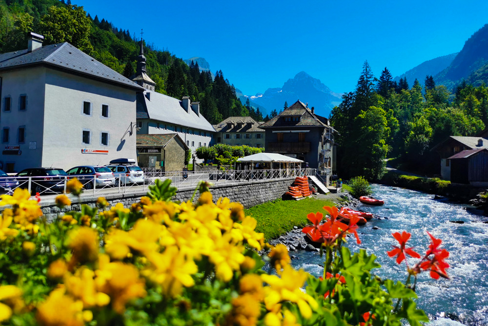 Scenic riverfront village with colorful flowers in the foreground, white buildings, and forested mountains under a clear blue sky.