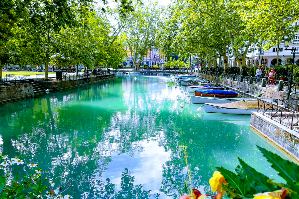 Turquoise canal lined with moored boats and a tree-filled promenade on both sides.