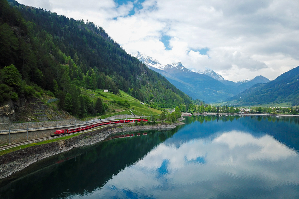 Red passenger train winds along a lakeside railway beside green hills and snow-capped mountains beyond the lake's edge.
