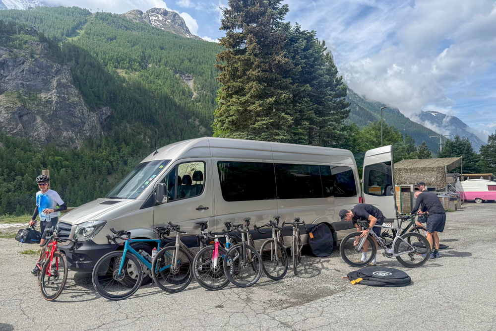 Group of cyclists prepping bikes beside a gray van in a mountain valley with pine trees and rugged cliffs behind them.
