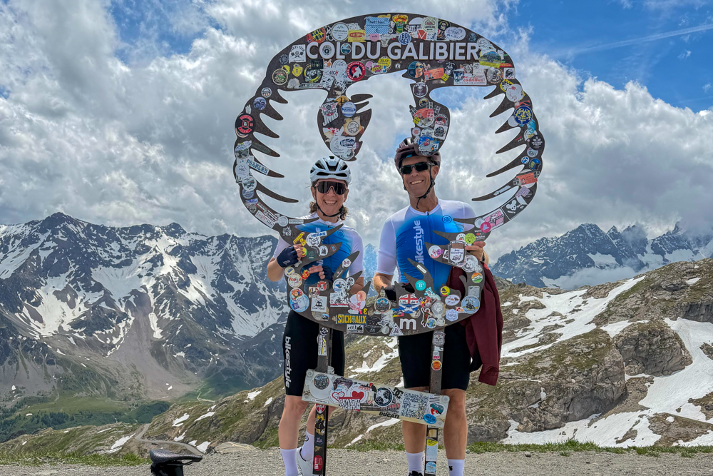 Two cyclists smiling beside a giant sticker-covered frame reading 'Col du Galibier' with snowy alpine mountains in the background on a sunny day.