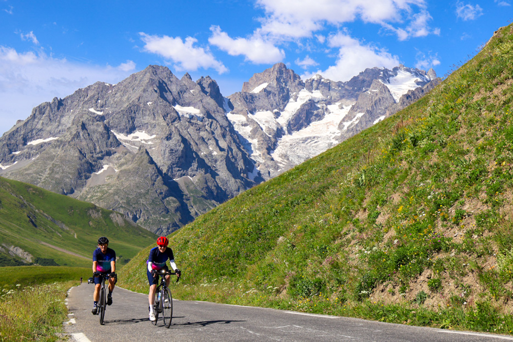 Two cyclists ride along a winding mountain road with snow-capped peaks in the background and green hills on either side.