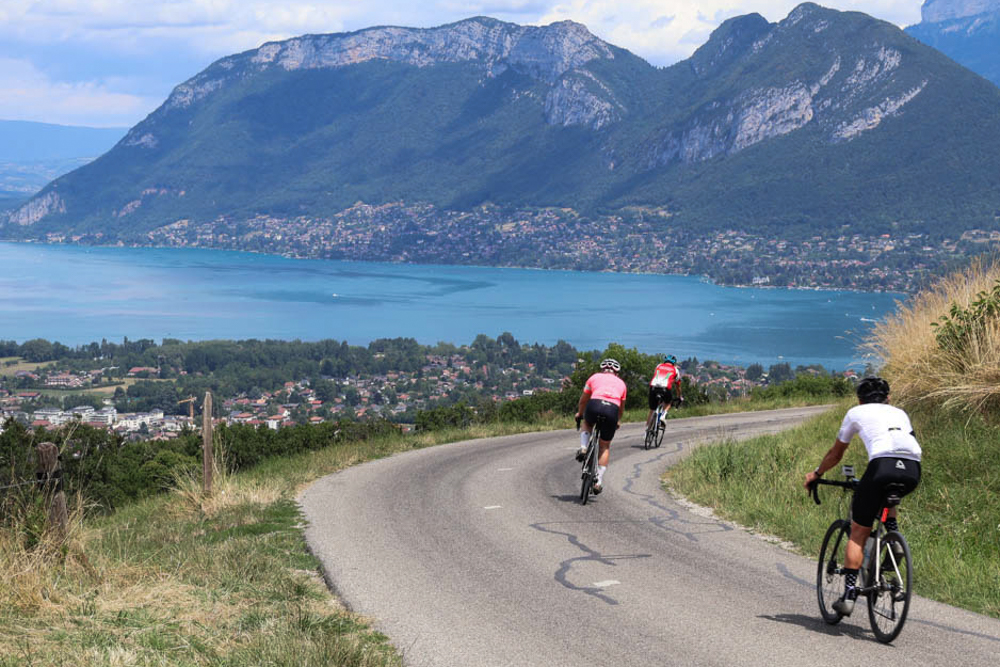 Three cyclists ride along a winding hillside road with a lake Anncey and town below, backed by tall mountains and a cloudy sky.