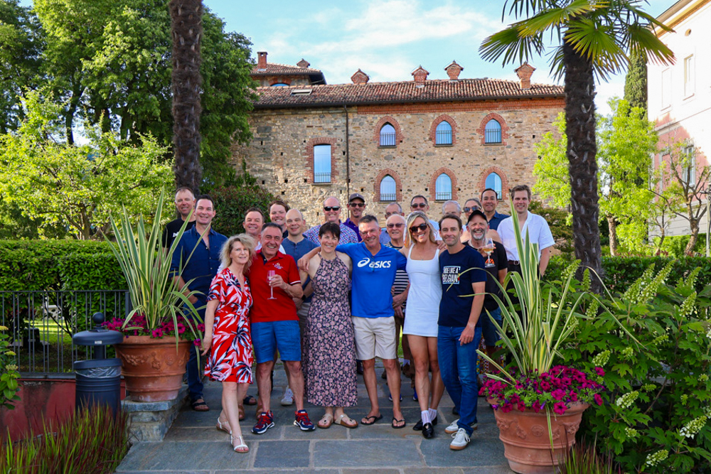 Group of smiling adults posing for a photo in a sunny garden courtyard with potted plants and a historic brick building in the background