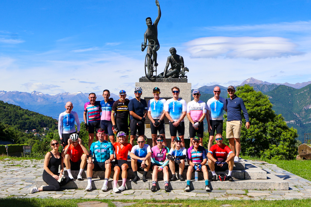 Group of cyclists posing for a photo in front of a statue on a stone pedestal, with mountains behind them.