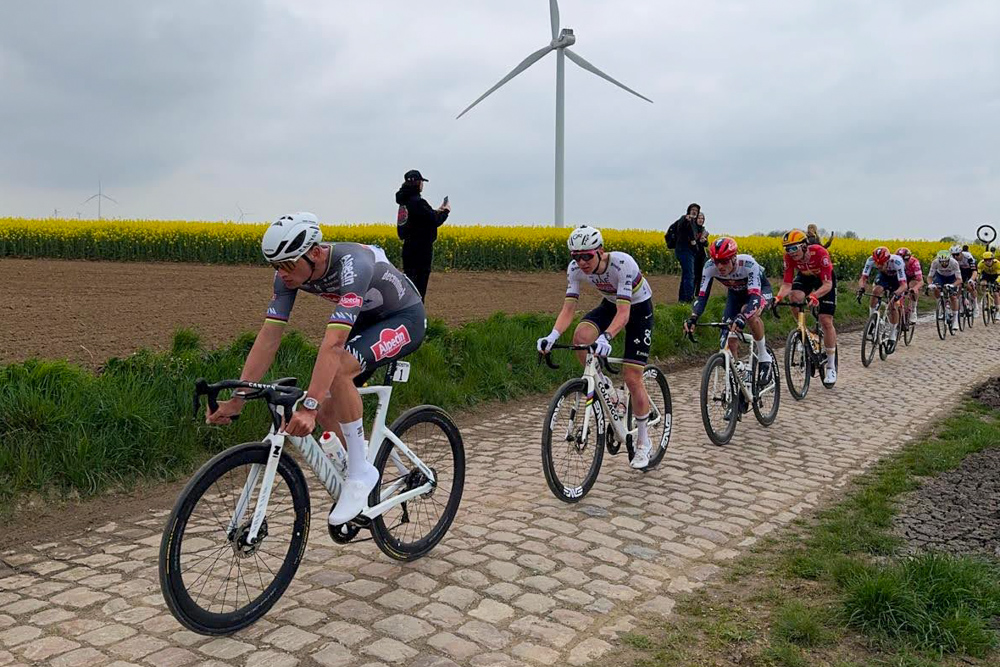 Professional cyclists riding a cobbled road past fields and a wind turbine.