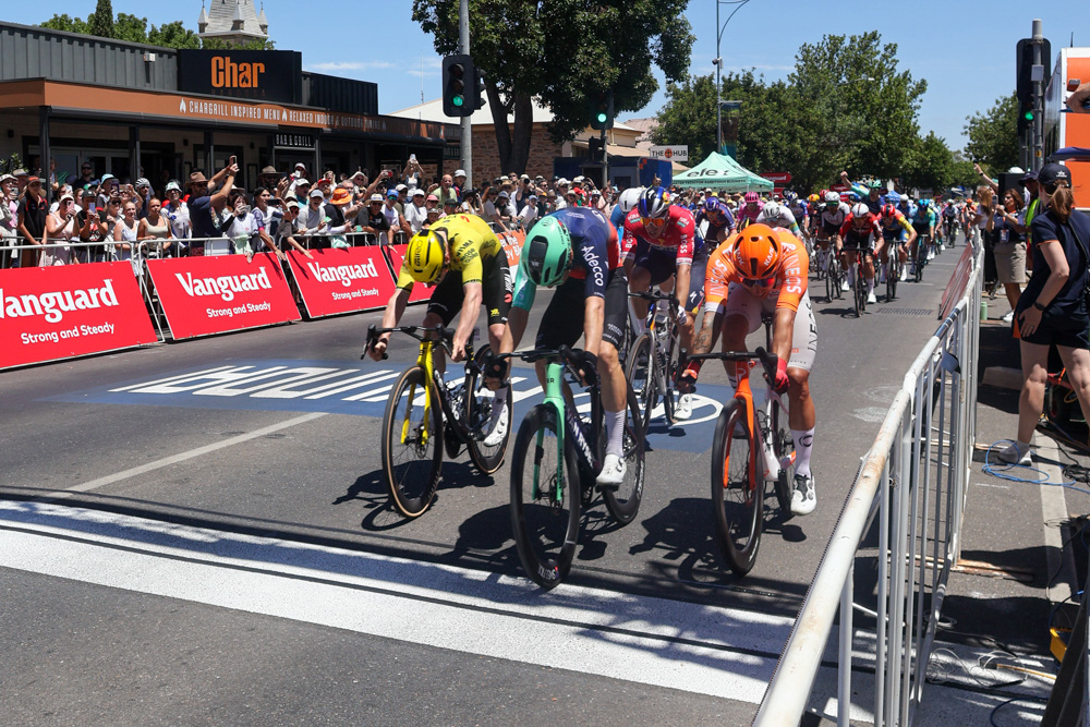 Group of professional cyclists sprinting on a city street as crowds cheer behind red Vanguard barriers at a race finish line.