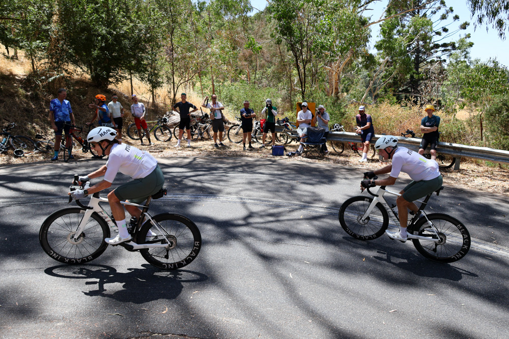 Two cyclists in white jerseys race on a sunlit road as a group of spectators watches from the roadside under trees.