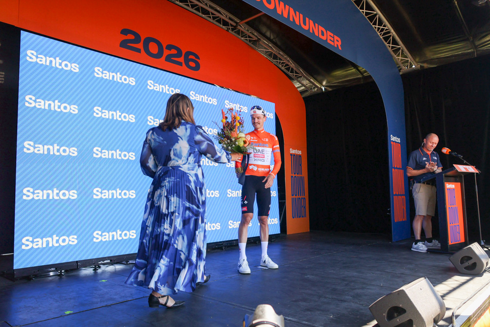 Cyclist in a red jersey accepts a bouquet from a woman in a blue dress on a stage with a blue Santos backdrop and '2026' signage.